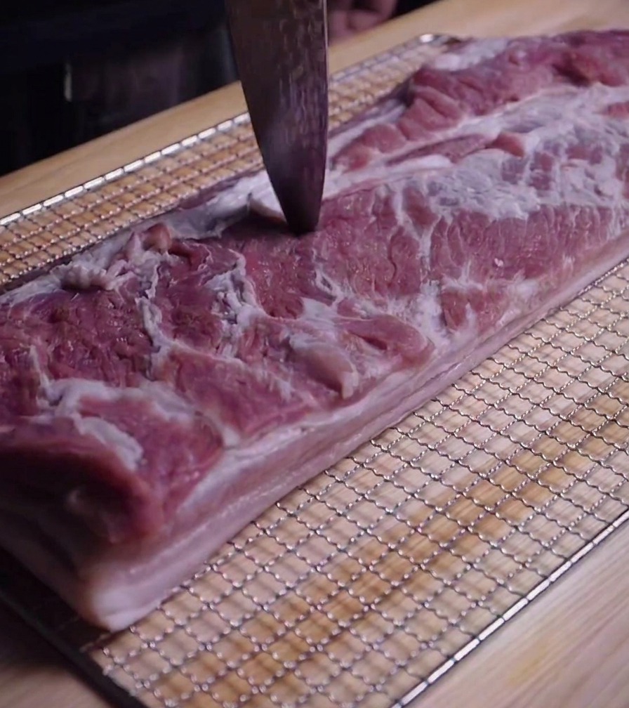 Close-up of a knife tip being used to pierce small holes into the surface of raw pork belly meat.