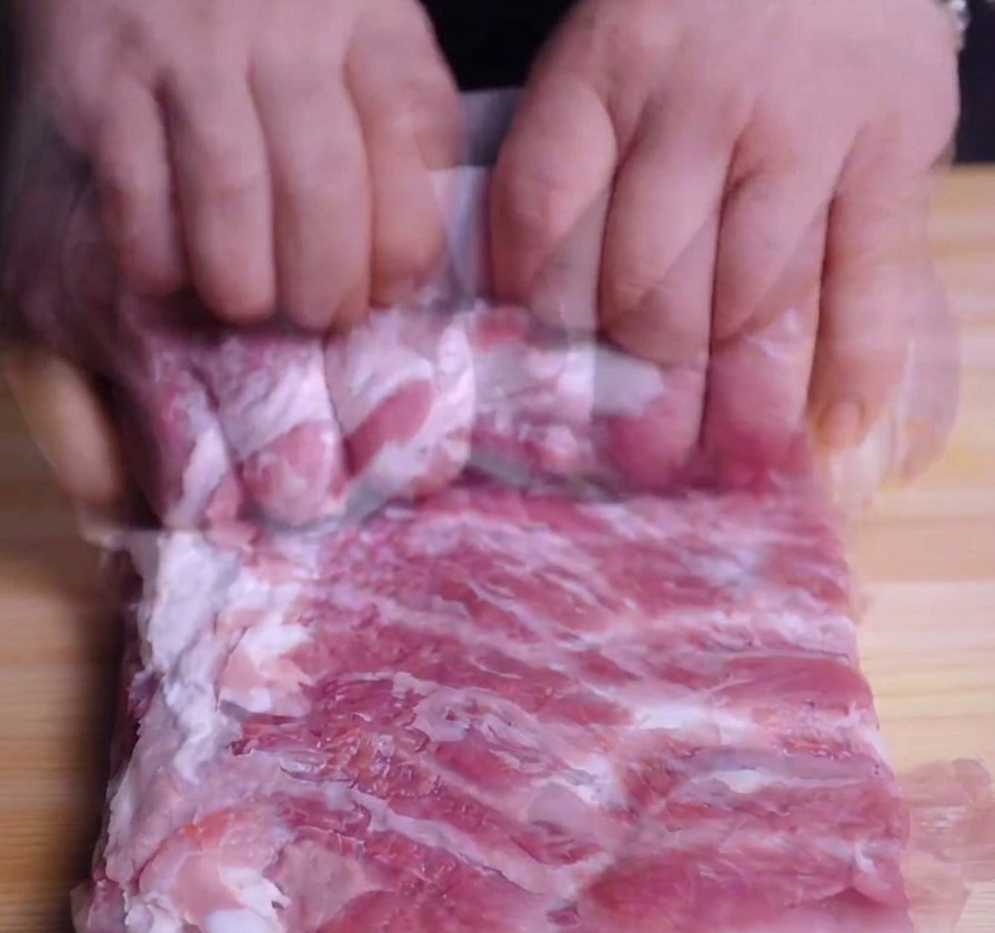 A persons hands tightly rolling a slab of raw pork belly into a cylinder on a wooden cutting board.