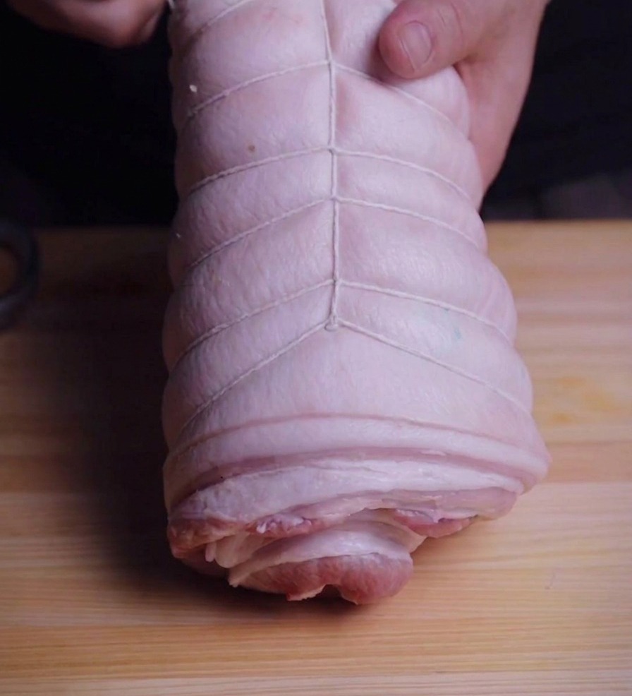 Hands holding a tightly rolled pork belly cylinder while wrapping it with white kitchen twine on a wooden board.