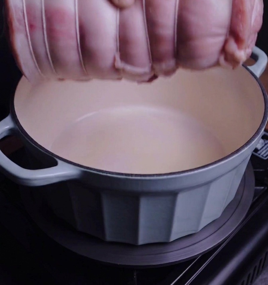 A tied pork belly roll being lowered by hand into a light blue ceramic-coated enamel Dutch oven.