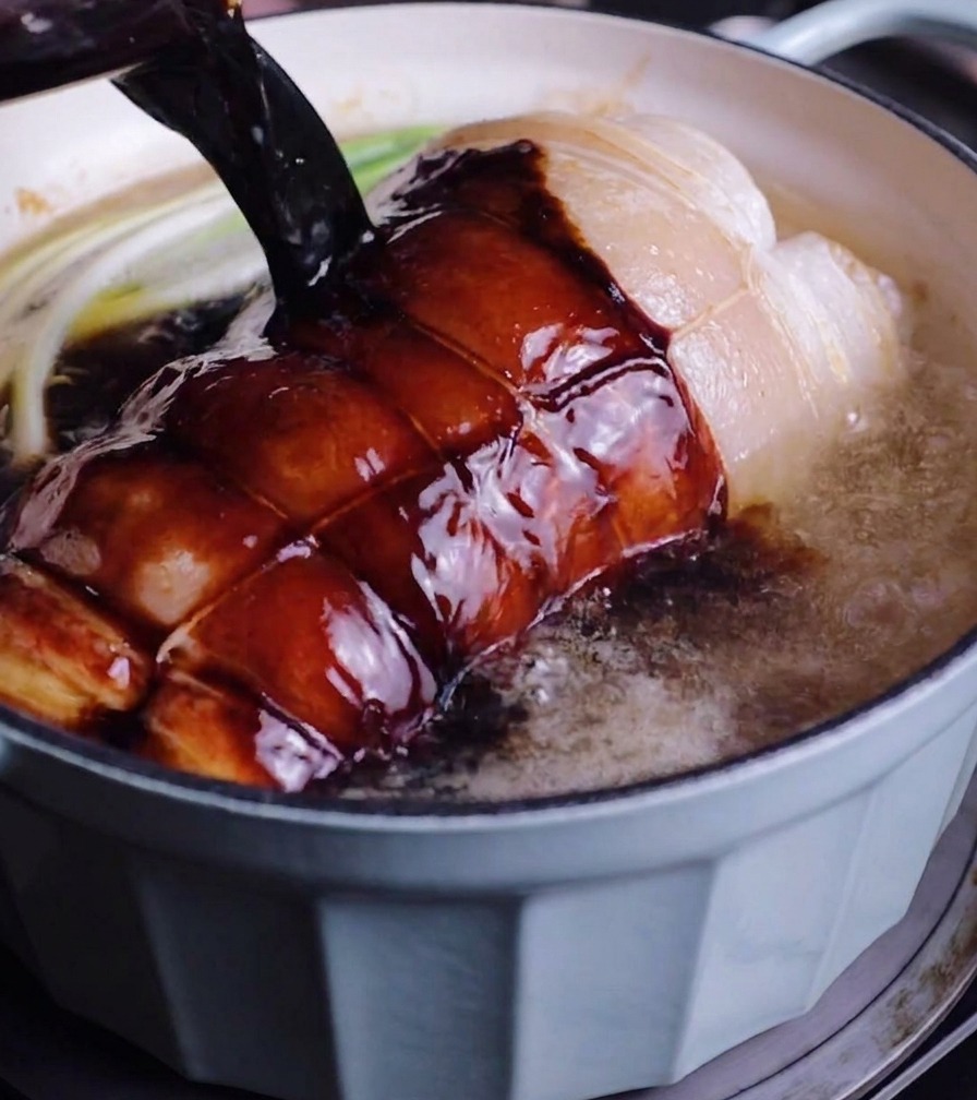 Dark braising liquid being poured from a glass measuring cup over a tied pork roll and green scallions inside a blue pot.