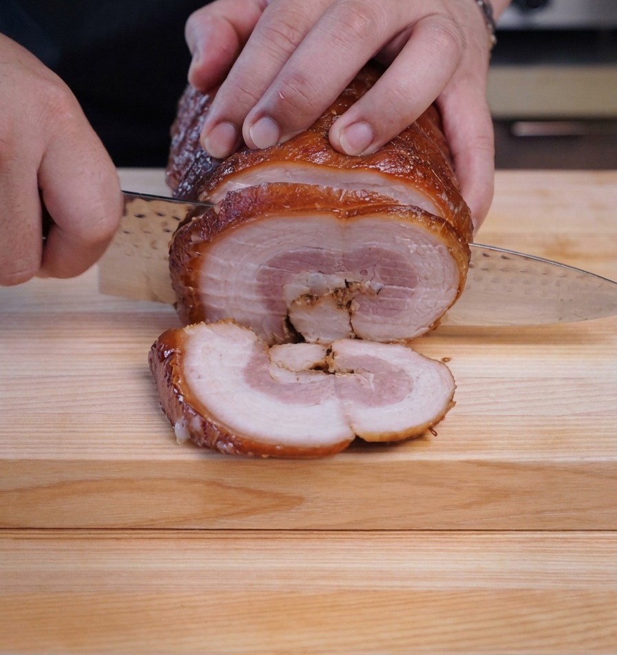 A chef using a sharp knife to cut thick, round slices from a chilled, braised chashu pork log.