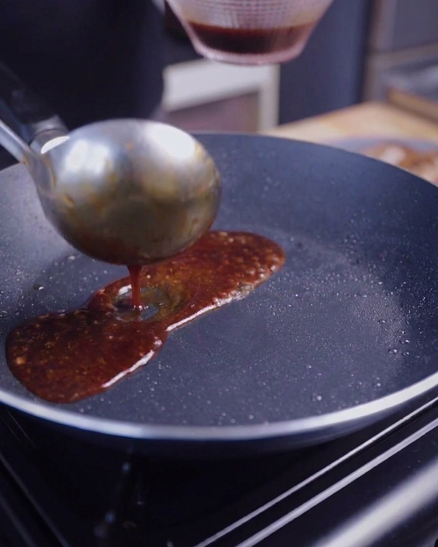 A metal ladle pouring a dark brown, glistening sauce into a pan containing seared pork belly slices.