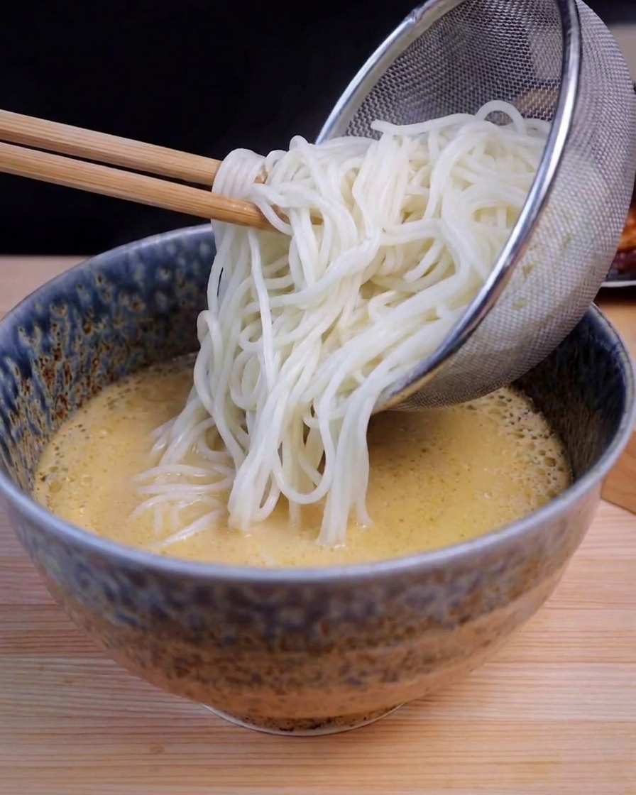 A stainless steel mesh strainer pouring long white noodles into a blue ceramic bowl containing yellow ramen broth.