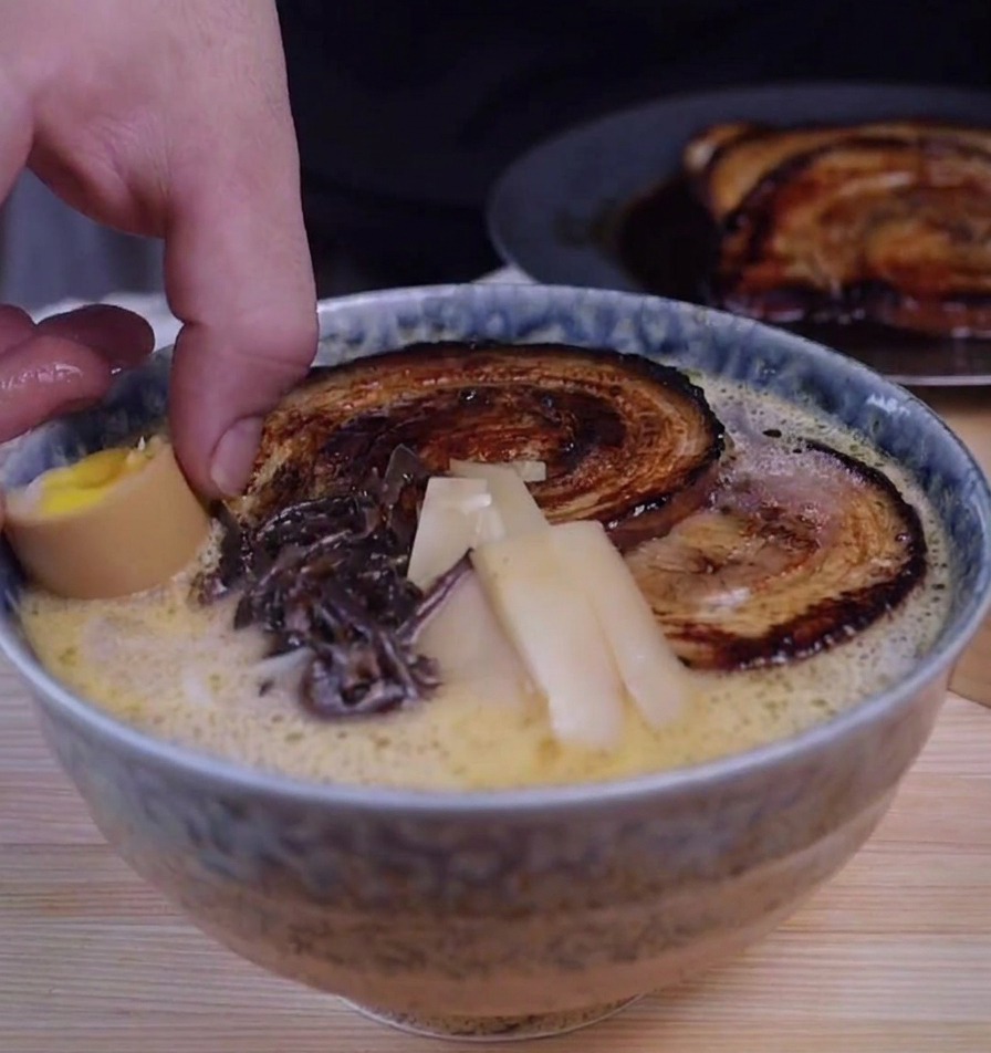 A hand placing a halved soft-boiled egg into a bowl of tonkotsu ramen already garnished with charred chashu slices, wood ear mushrooms, and bamboo shoots.