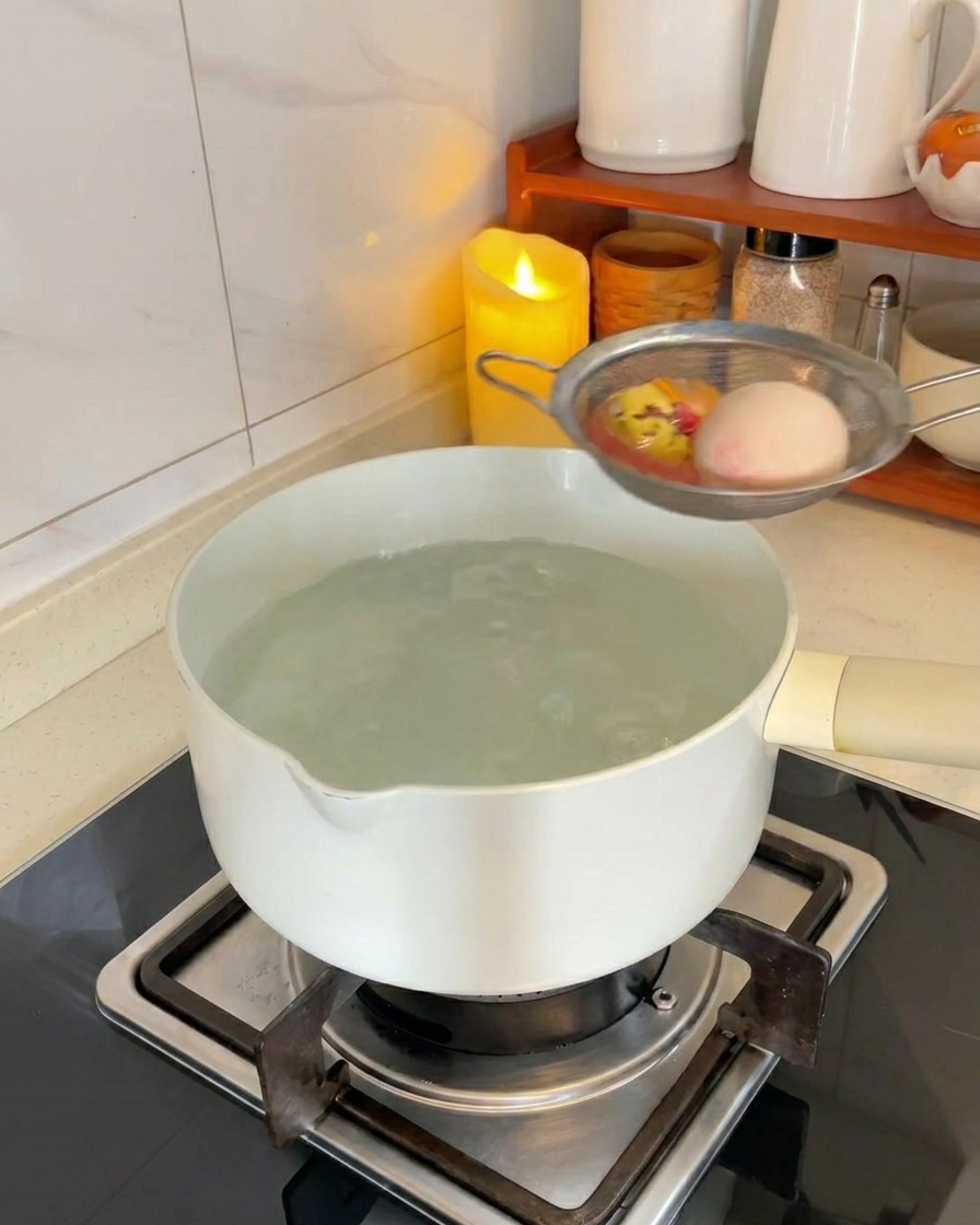 Two white eggs in a metal mesh strainer being lowered into a white pot of boiling water on a kitchen stove.