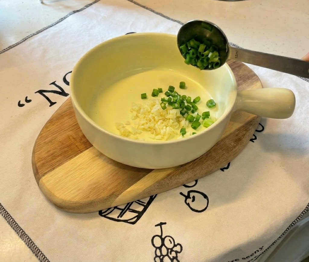 A small yellow ceramic bowl resting on a wooden board, containing mounds of finely minced garlic and sliced green onions.