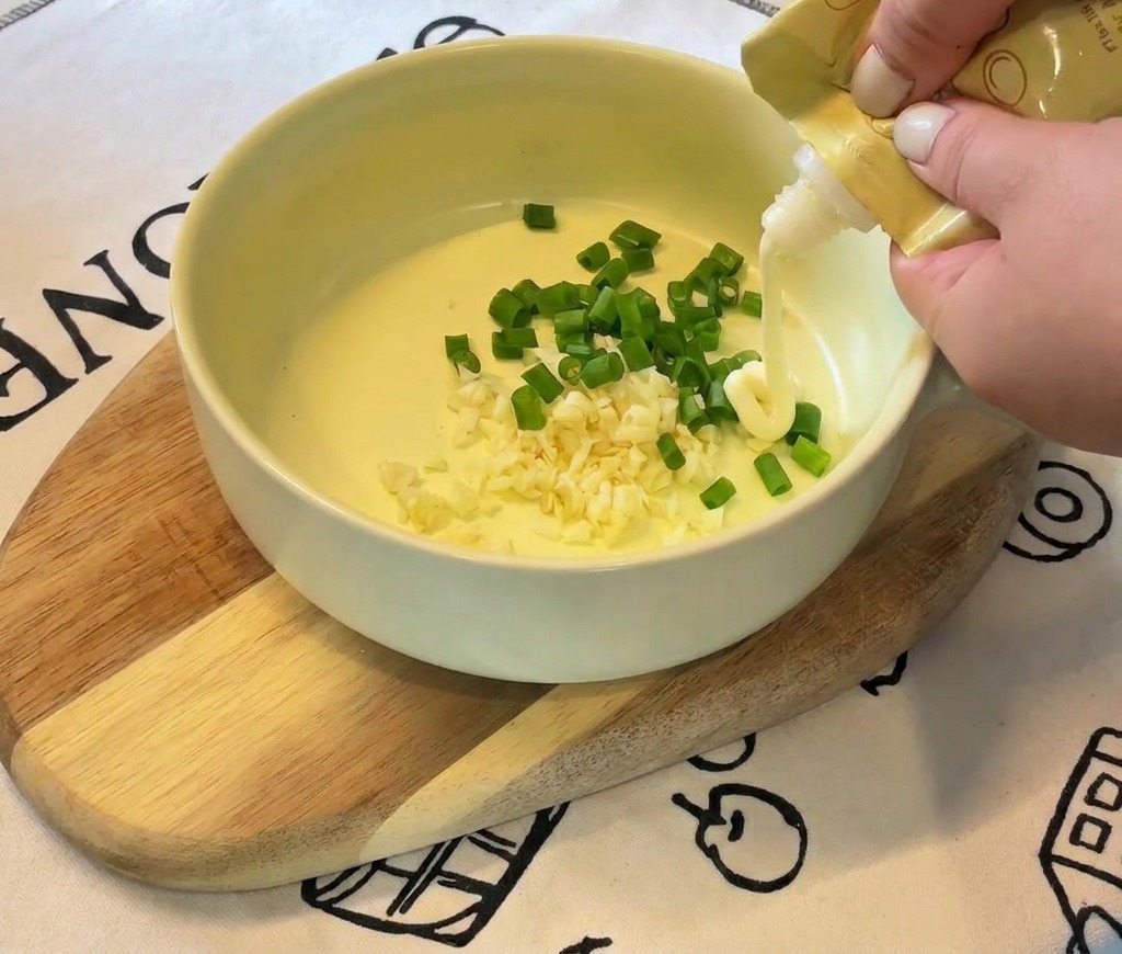A persons hands squeezing white mayonnaise from a yellow tube into a bowl containing garlic and green onions.