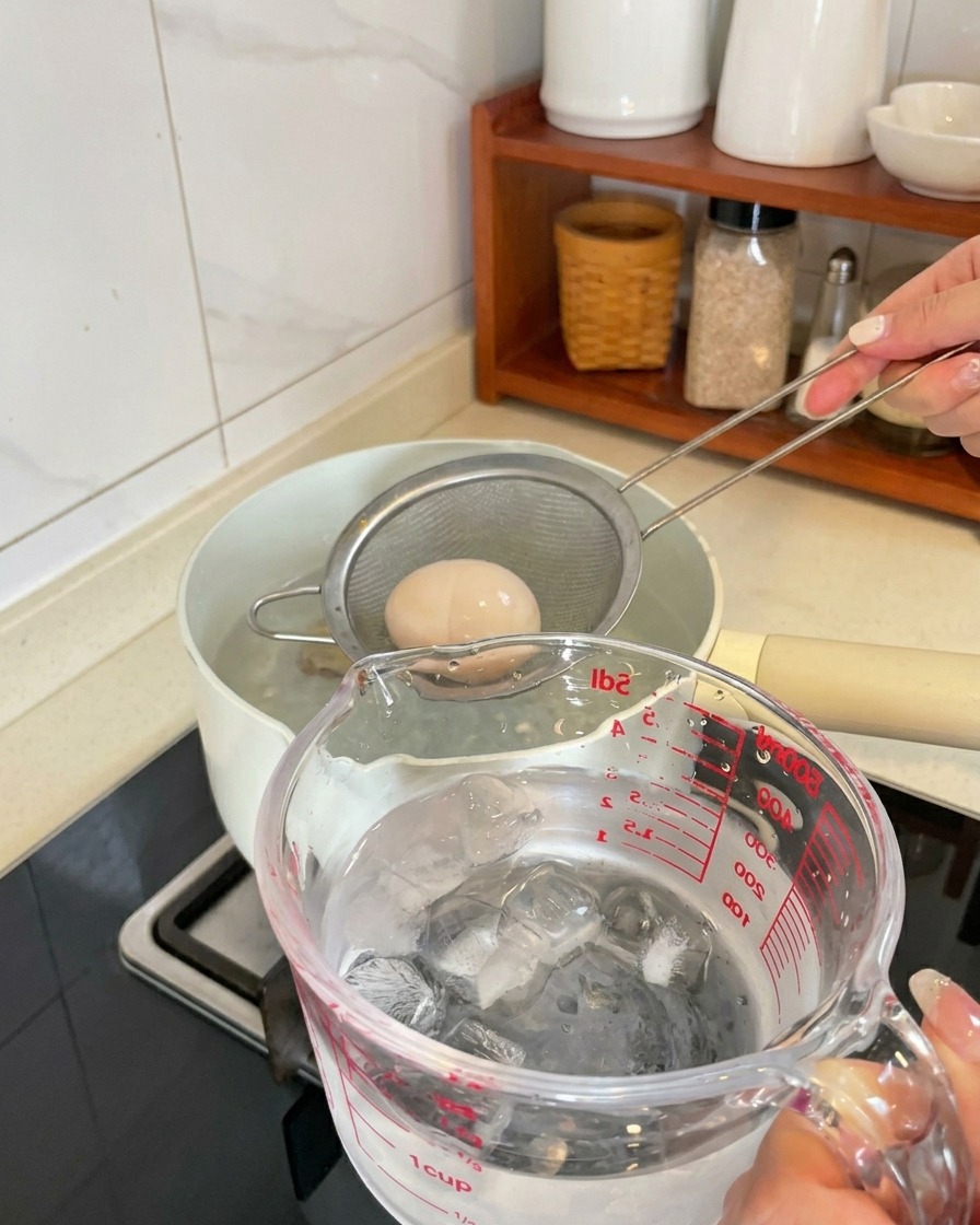A boiled egg being lifted from a pot in a mesh strainer and placed into a measuring cup filled with ice cubes and water.