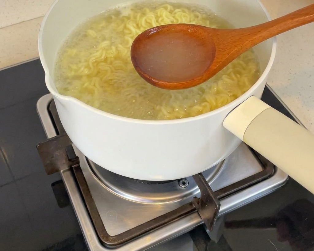 A wooden ladle lifting clear, steaming noodle cooking water from a white pot over a stovetop burner.