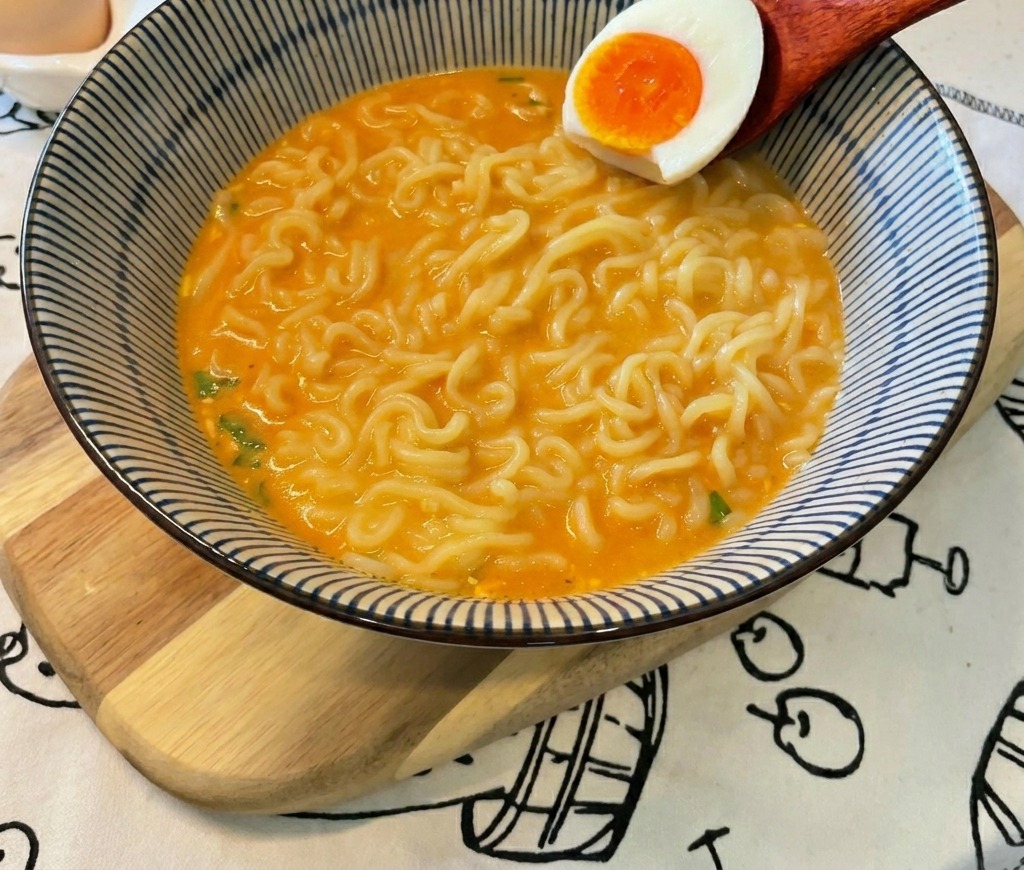 A wooden spoon lowers a soft-boiled egg half with a vibrant orange yolk into a bowl of ramen noodles and orange broth.