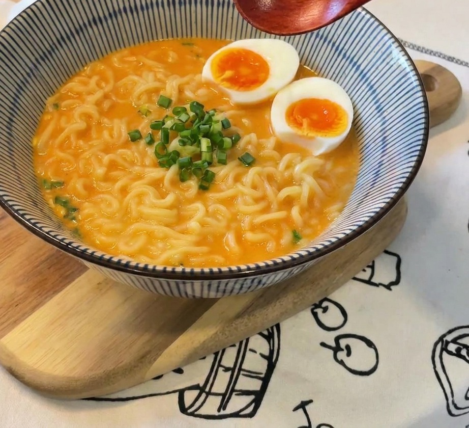 A bowl of ramen topped with eggs and a fresh pile of sliced green onions in the center, with a wooden spoon held above the dish.