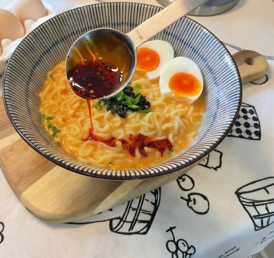 Vibrant red chili oil is poured from a metal measuring spoon onto a completed bowl of ramen with eggs, onions, and seaweed.