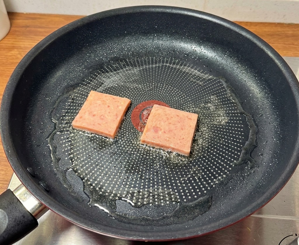 Slices of pink luncheon meat being seared in a black frying pan until they show golden-brown charred marks.