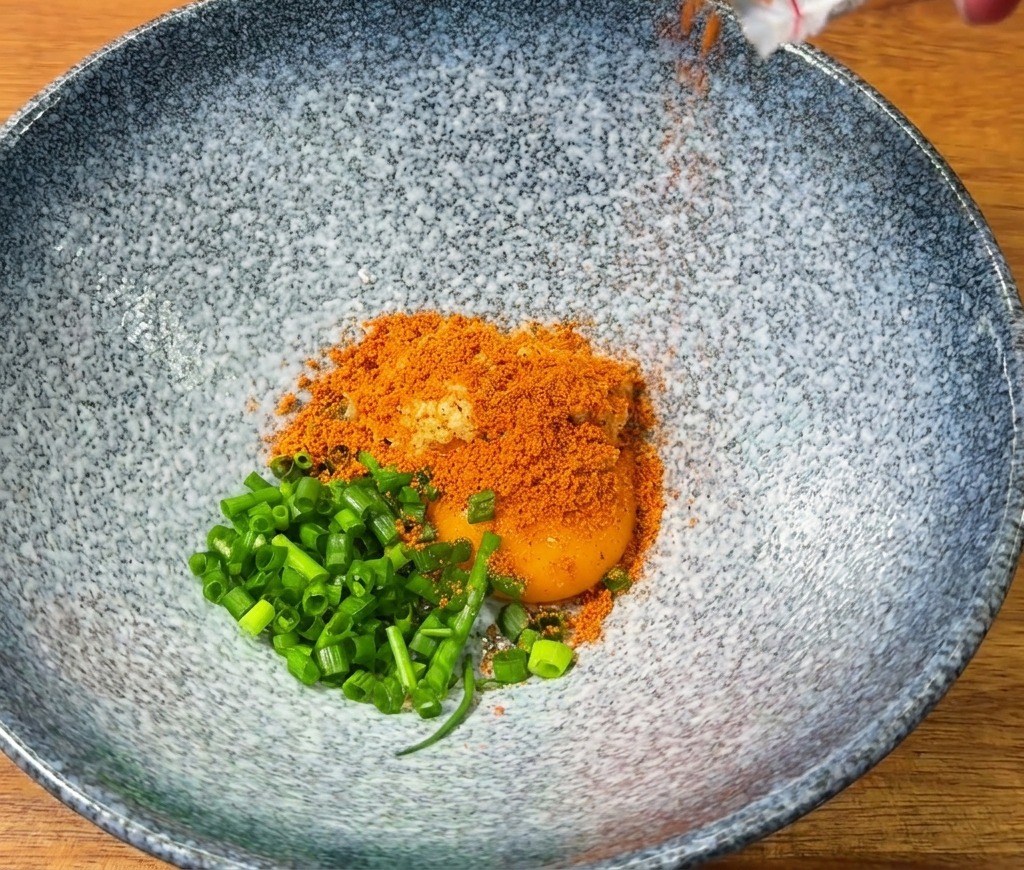 Red spicy ramen seasoning being poured into a blue bowl containing an egg yolk and fresh green onions.