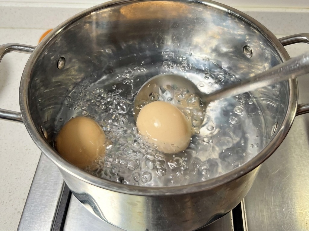 A stainless steel slotted spoon lifting two brown eggs out of a pot of boiling water.