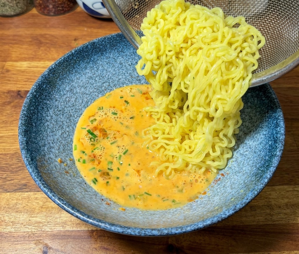 Cooked yellow ramen noodles being poured from a stainless steel mesh strainer into a blue bowl of orange-tinted creamy broth.