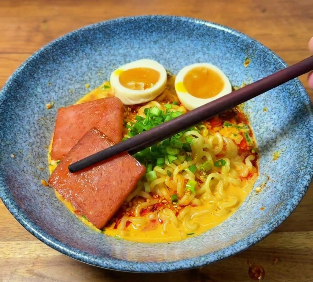 Chopsticks placing a piece of pan-fried luncheon meat into a finished ramen bowl already garnished with eggs and chili oil.