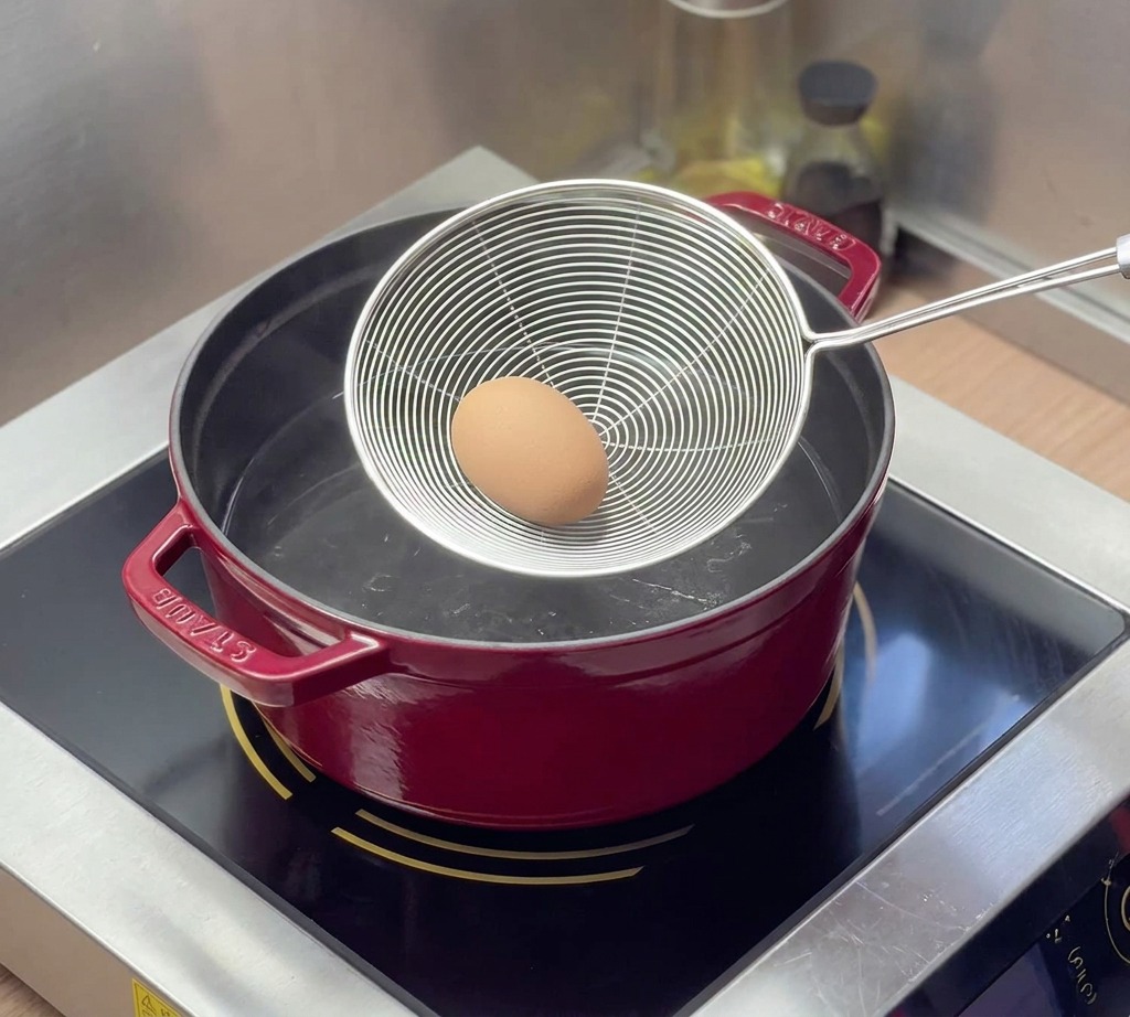 A brown egg rests in a stainless steel mesh spider strainer as it is being lowered into a red Staub pot filled with boiling water.