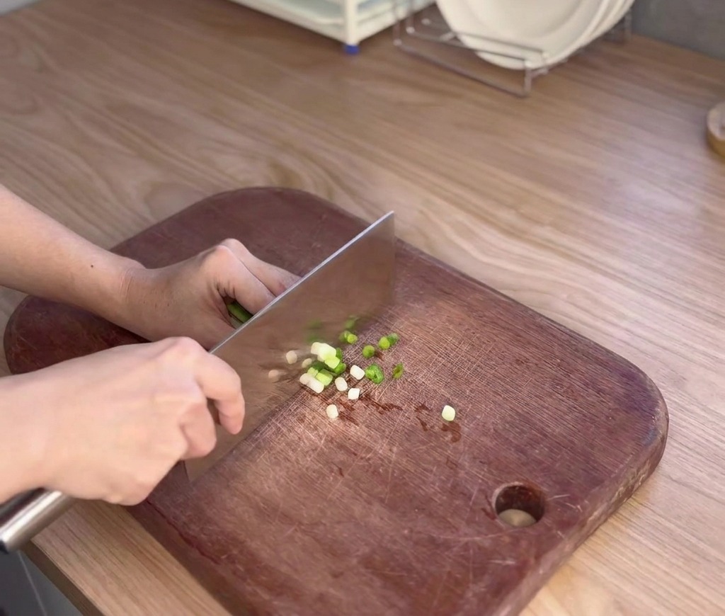 A person uses a large kitchen knife to finely slice fresh scallions into small rounds on a dark wooden cutting board.