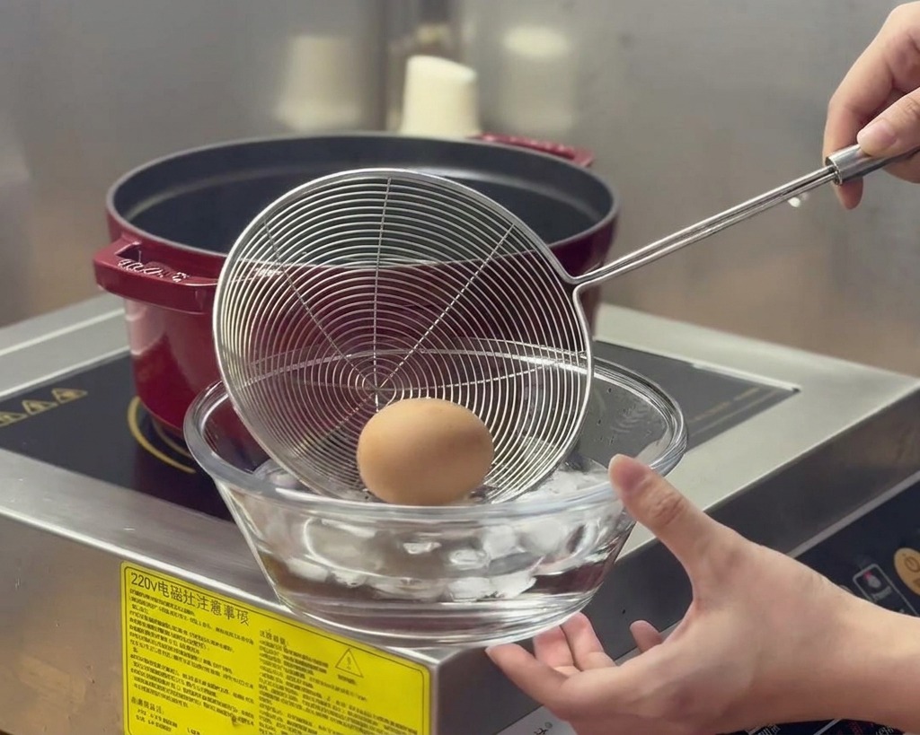 A boiled egg is moved via a mesh strainer from a steaming red pot into a glass bowl filled with water and ice cubes.