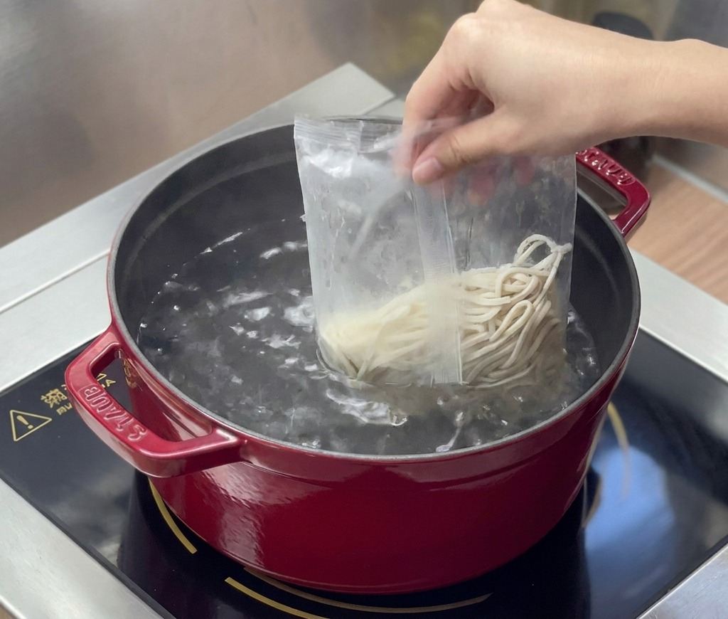 Fresh ramen noodles are poured from a clear plastic bag into a red pot of boiling water.