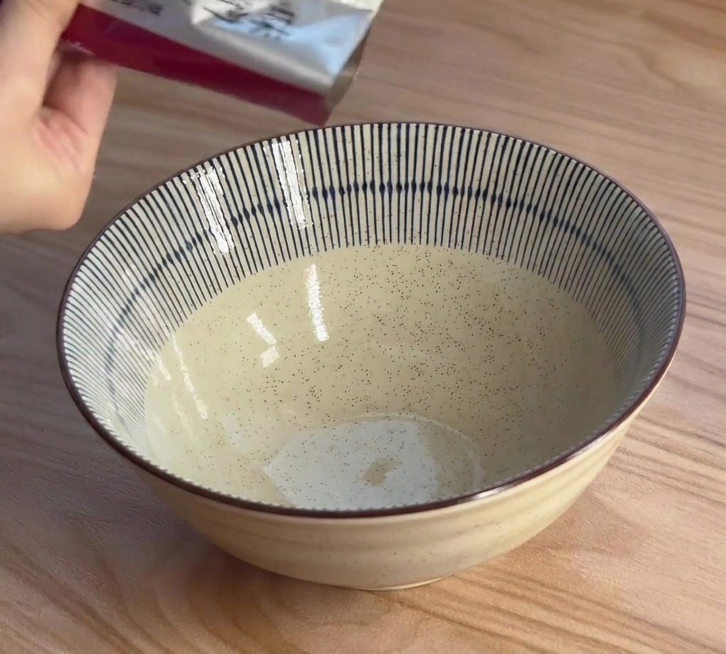 A hand squeezes thick miso ramen seasoning paste from a silver foil packet into a blue and white striped ceramic bowl.