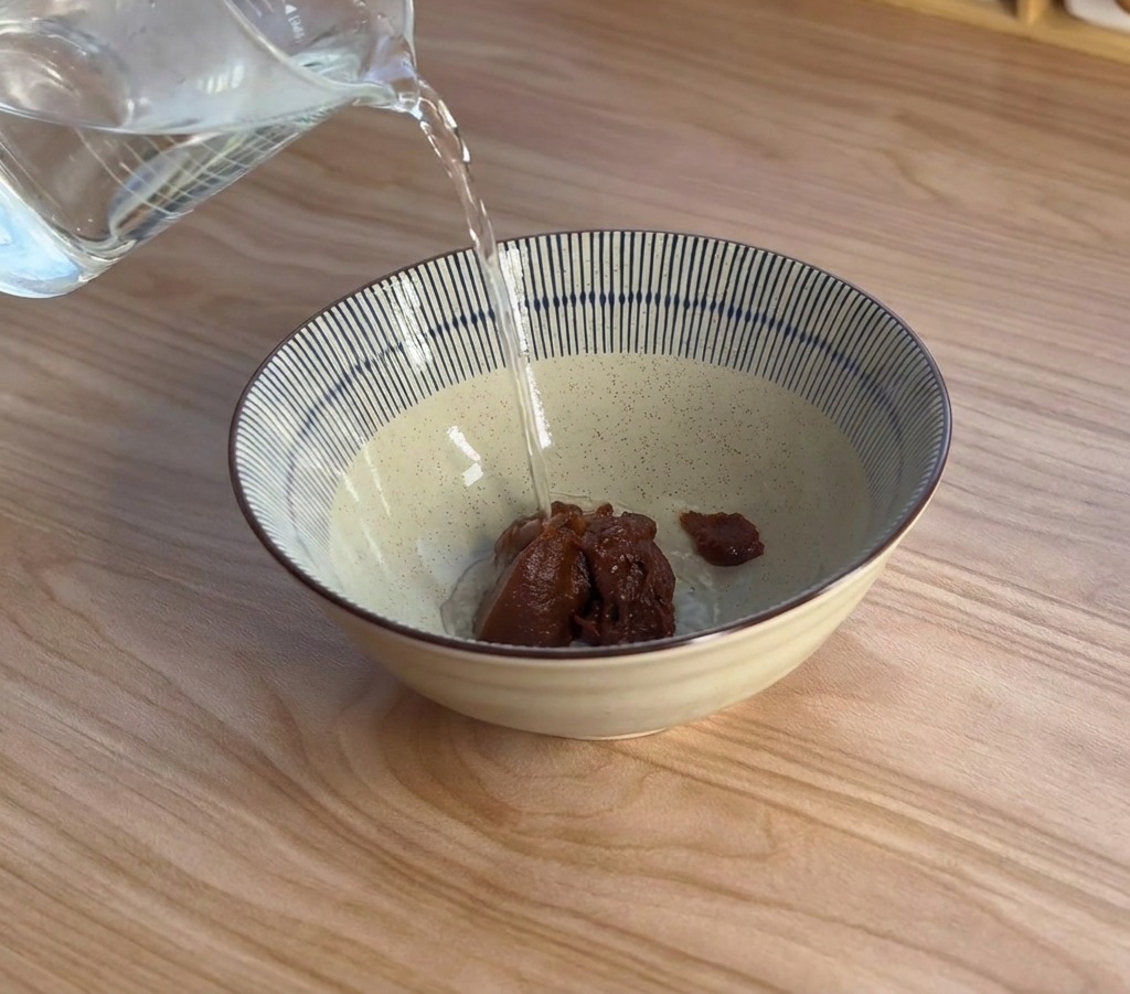 Steam rising as boiling water is poured from a measuring cup into a ceramic bowl containing a scoop of dark miso paste.