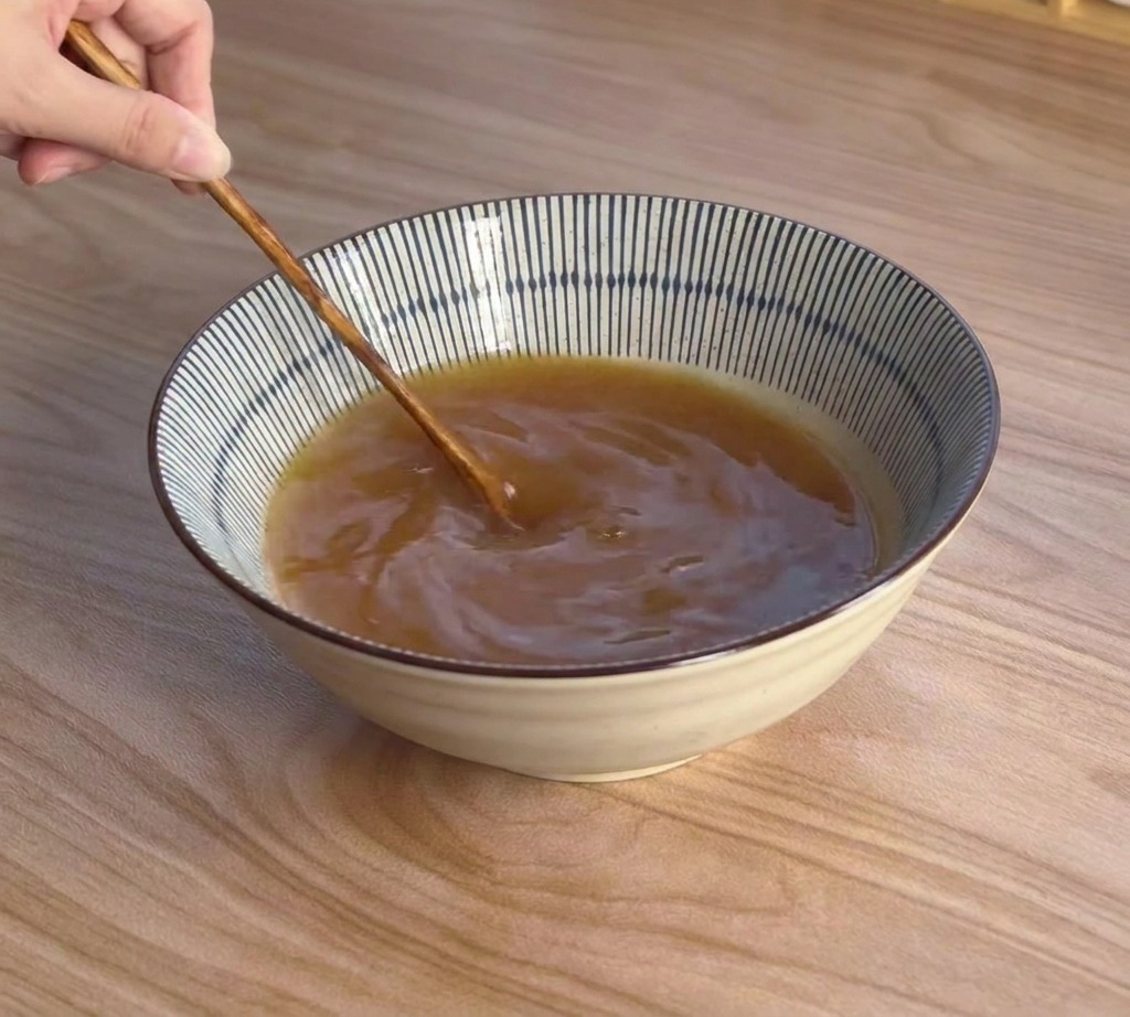 A hand using a long-handled wooden spoon to stir a rich, brownish miso broth in a blue and white striped bowl.