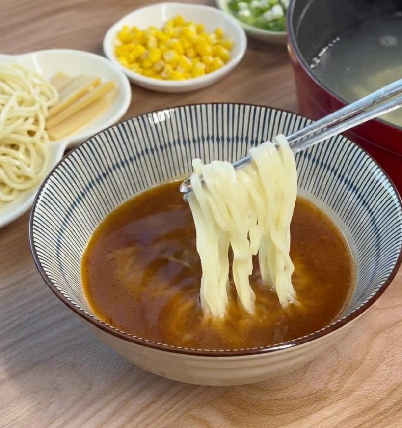 Cooked ramen noodles being added to and swirled within a bowl of brown miso broth using chopsticks.