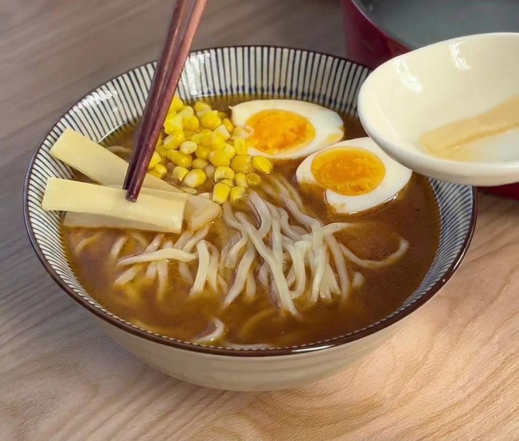 Dark wooden chopsticks placing thin, light-colored bamboo shoot slices into a bowl of ramen already topped with egg and corn.