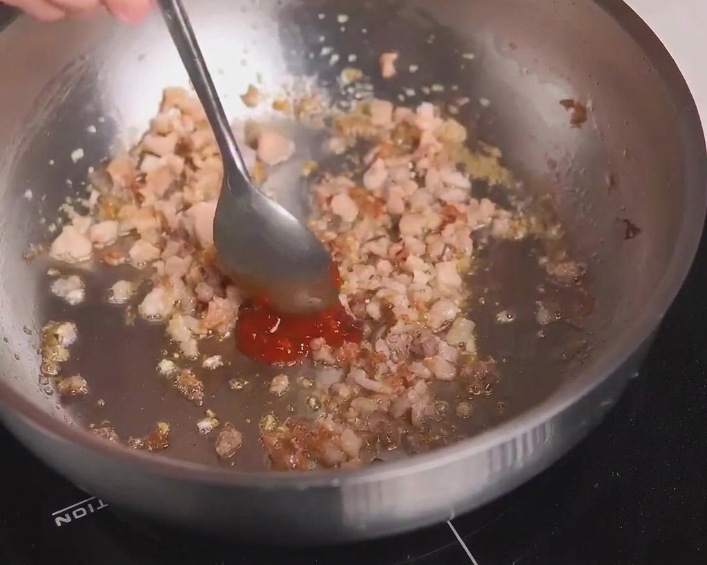 A metal spoon adding a dollop of red spicy bean paste to a pan containing browned ground meat.