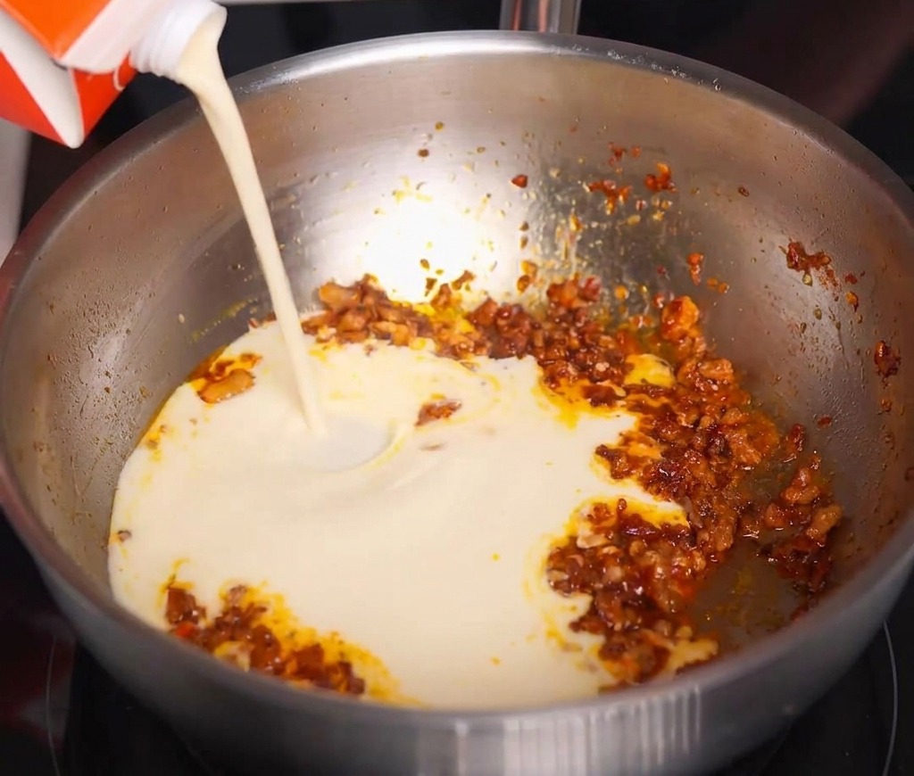 Creamy white soy milk being poured from a red and white carton into a pan of spicy sautéed meat.