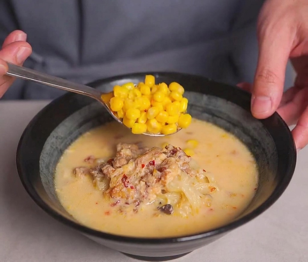 A metal spoon depositing a heap of golden sweet corn kernels onto the surface of a bowl of creamy ramen.