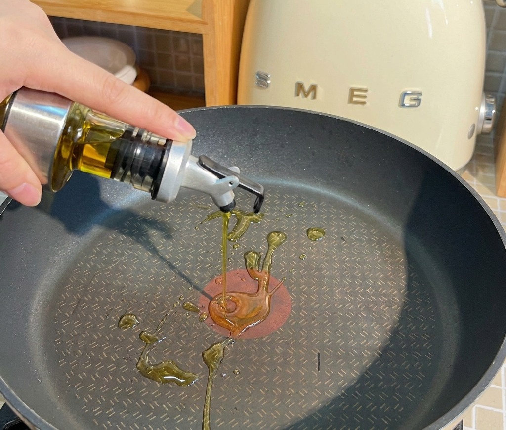 Cooking oil being poured from a glass dispenser into a black non-stick frying pan on a kitchen counter.