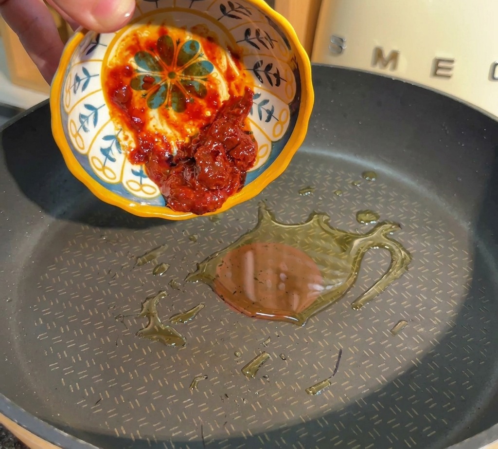 A hand pouring red spicy bean paste from a small patterned bowl into a frying pan with hot oil.