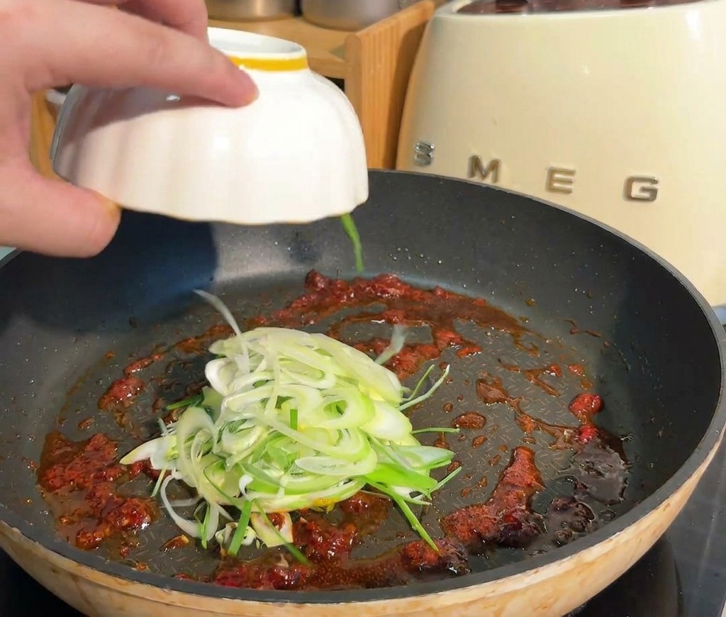 Sliced green onions being added from a white bowl into a pan containing a red spicy sauce base.