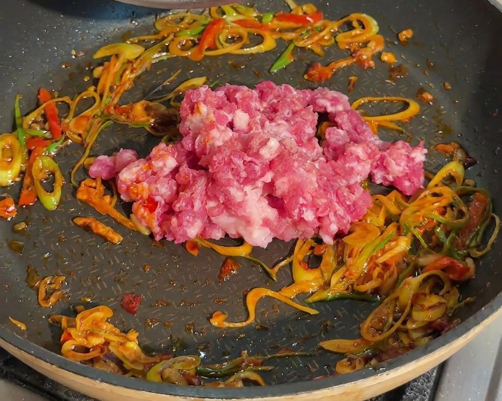 Raw minced pork being placed into a frying pan already containing sautéed green onions and red bean paste.