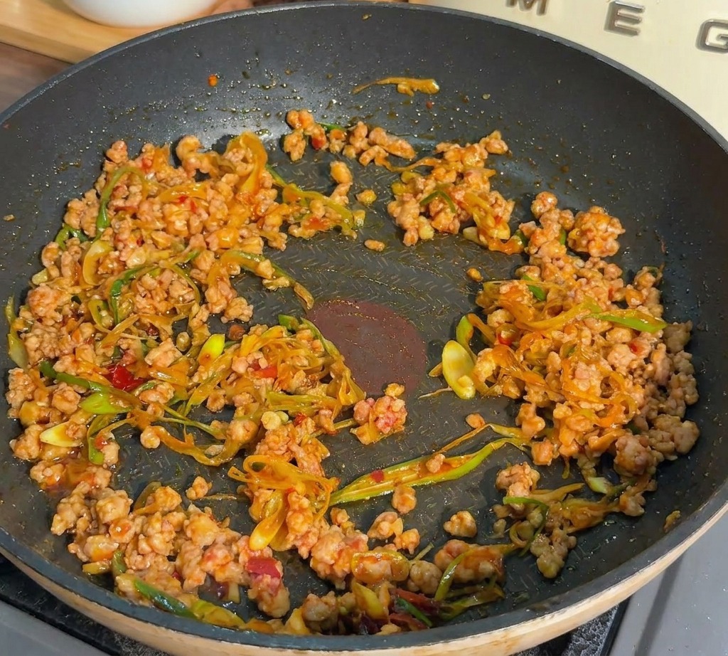 Minced pork and green onions being stir-fried in a dark pan until the meat appears browned and cooked.