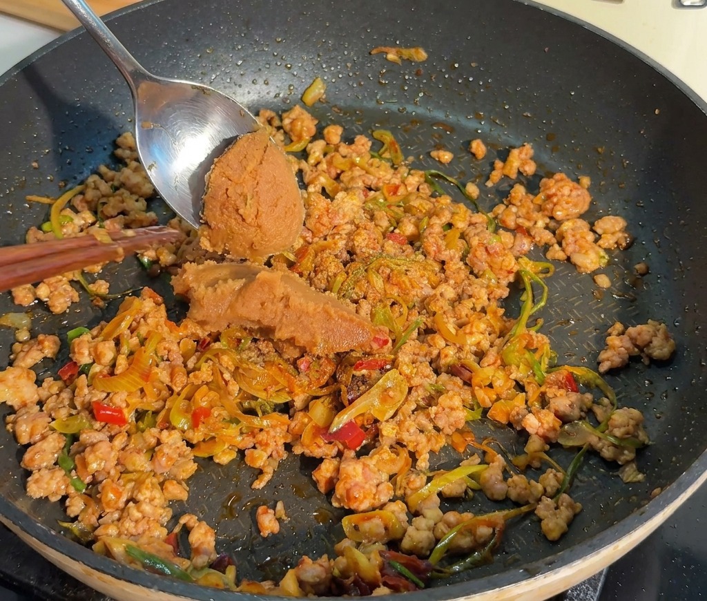 A metal spoon adding a portion of thick brown miso paste to a pan of cooked minced meat.