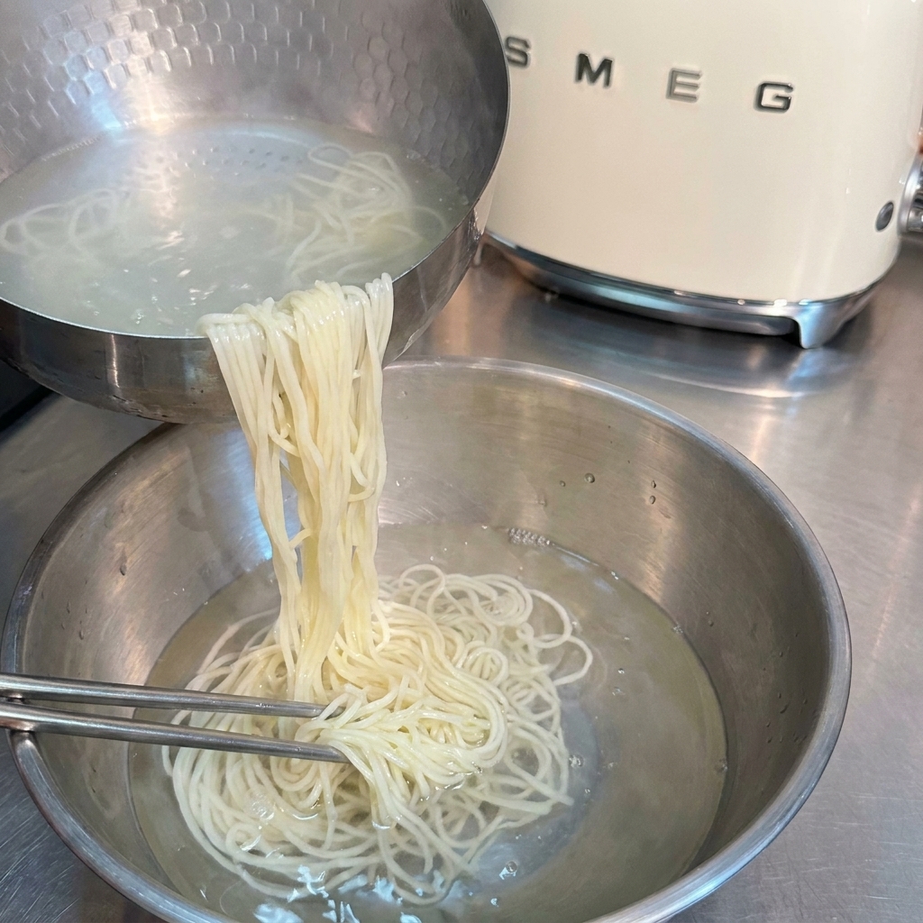 Cooked ramen noodles being rinsed and agitated with metal chopsticks in a stainless steel bowl of cold water.