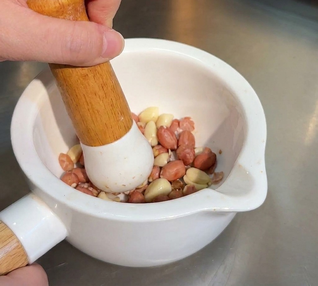 A person using a wooden pestle to crush roasted peanuts and sesame seeds in a white ceramic mortar.