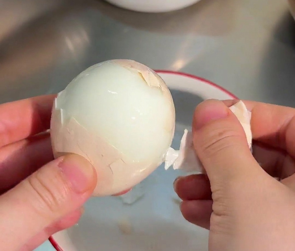 Hands carefully peeling the shell off a glistening, smooth white soft-boiled egg over a small bowl.
