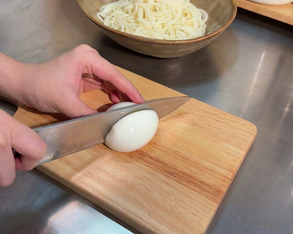 A sharp kitchen knife slicing through the middle of a soft boiled egg on a light colored wooden cutting board.