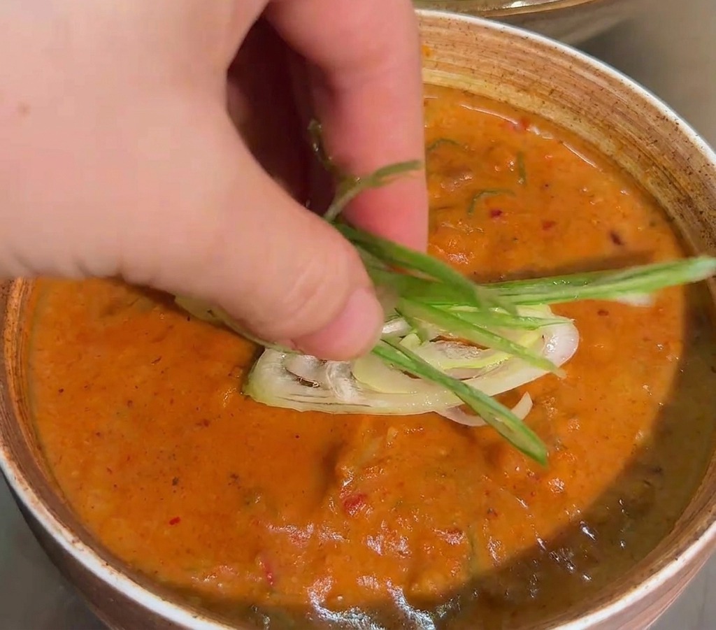 A hand placing a bundle of thin green onion strips onto the surface of a thick orange dipping sauce in a ceramic bowl.