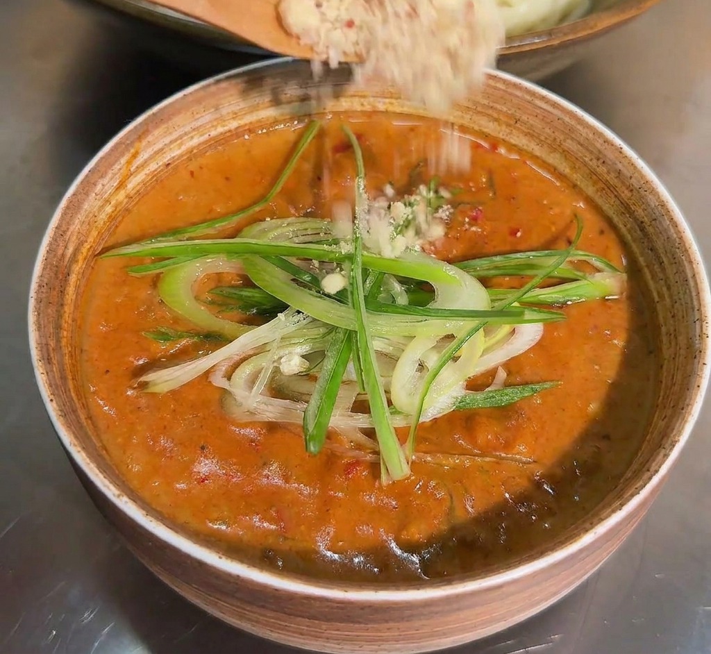 A wooden spoon sprinkling a light-colored ground nut and seed powder over a bowl of dipping sauce already garnished with green onions.