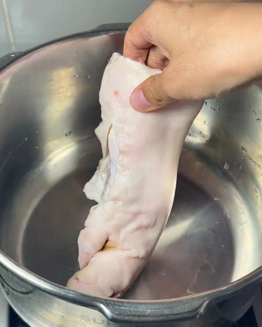 A hand placing a raw pigs trotter into a stainless steel pot alongside a large pork marrow bone and several chicken feet.