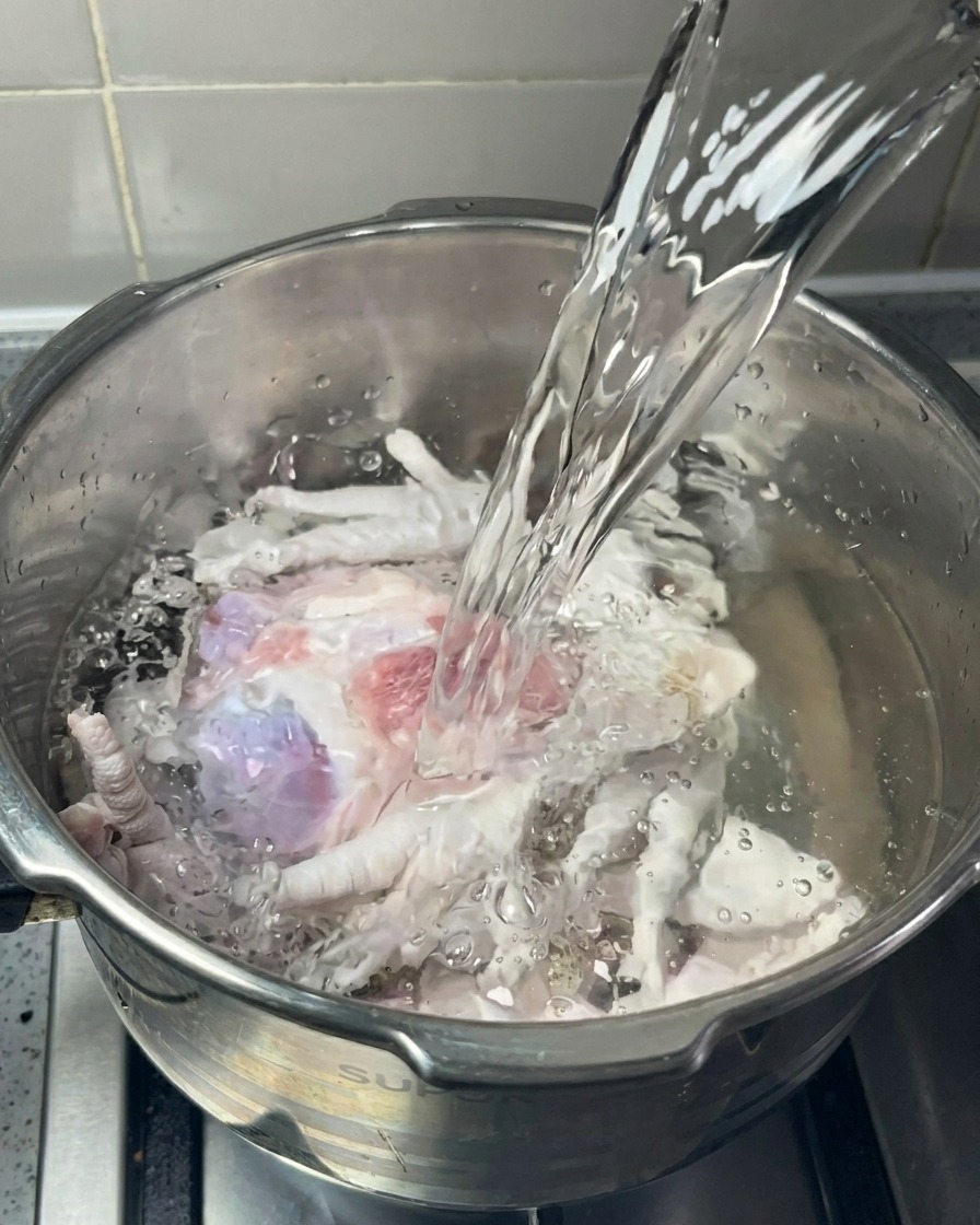 A stream of clear water being poured into a stainless steel pot filled with raw pork bones and chicken feet.
