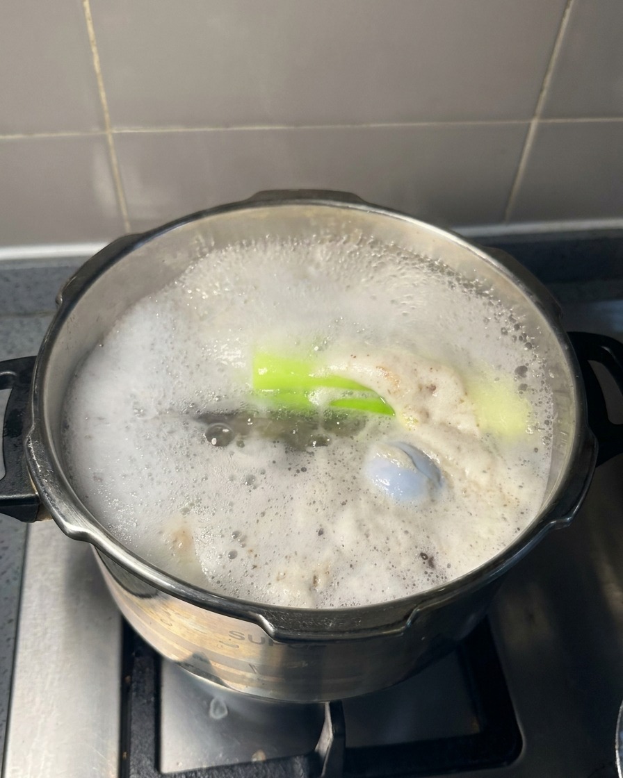 A large metal spoon lifting grey foam and bubbles from the surface of a boiling meat broth.