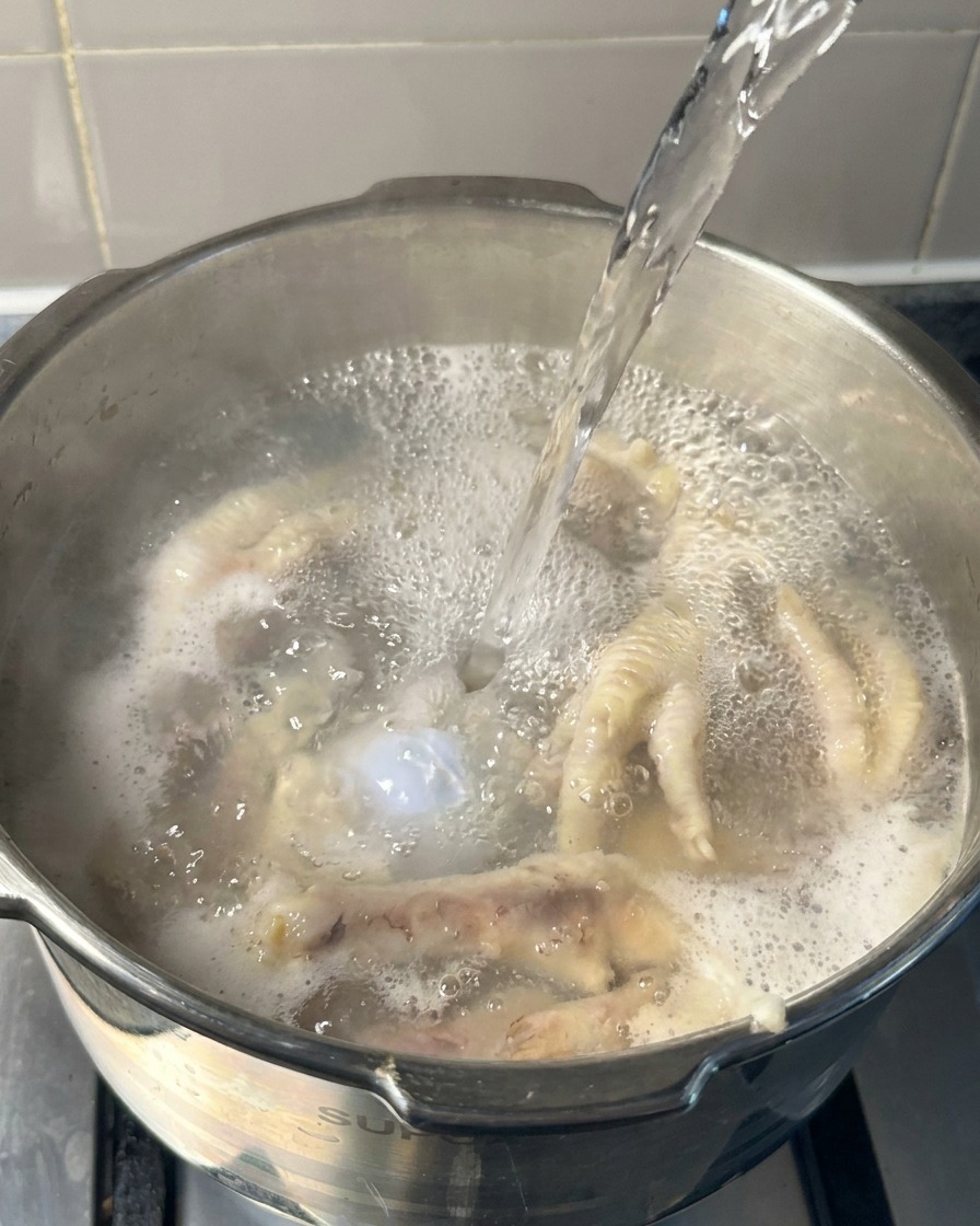 Boiling water being poured into a pot containing partially cooked chicken feet and pork bones.